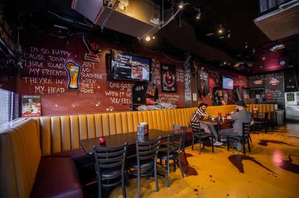 Spacious interior of a pizza and karaoke bar featuring long cushioned booths, chalkboard-style murals, mounted TVs, and casual dining tables.