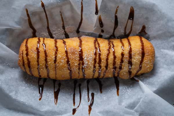Deep-fried pastry roll dusted with powdered sugar and drizzled with chocolate sauce, indulgent fair-style dessert on parchment paper.