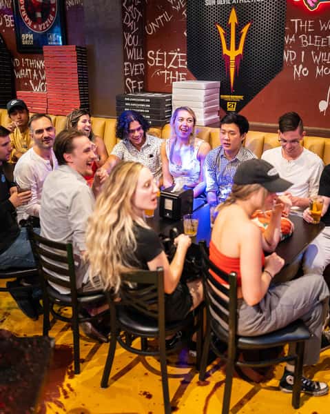 Large group of friends enjoying pizza and drinks inside a lively sports bar with booth seating, stacked pizza boxes, and energetic atmosphere.