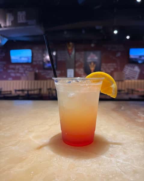 Refreshing layered cocktail with orange garnish served in a clear cup at a modern sports bar with cozy booth seating in the background.