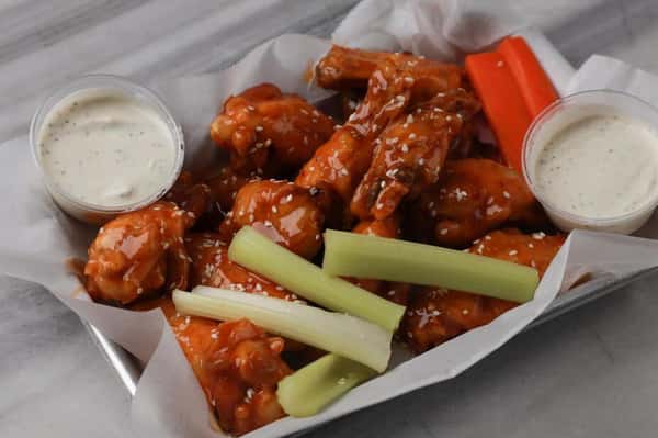 Buffalo chicken wings coated in tangy hot sauce and sprinkled with sesame seeds, served in a parchment-lined tray with crisp celery and carrot sticks and creamy ranch dipping sauce – classic spicy bar food perfect for a sports bar or casual American restaurant menu.