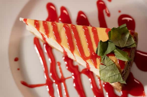Creamy New York-style cheesecake slice drizzled with strawberry sauce and topped with fresh mint, served on a white plate at a casual pizza restaurant.