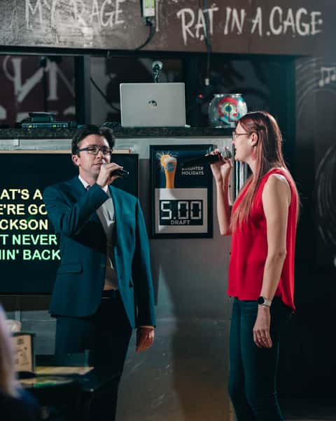 Two guests singing karaoke on stage at a lively pizza and sports bar, holding microphones under neon signs and chalkboard walls, creating a fun late-night entertainment atmosphere.