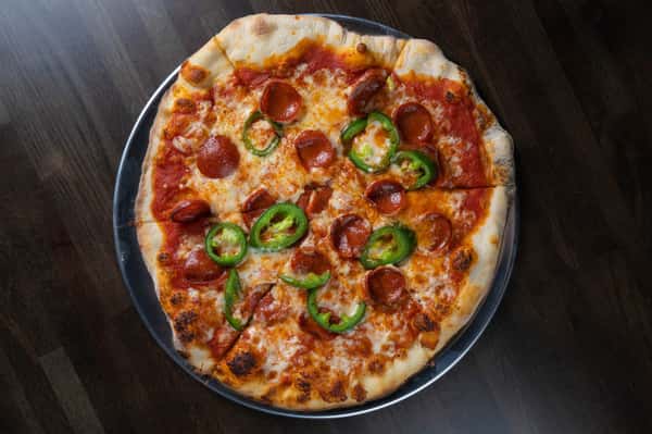 Wood-fired style pizza topped with pepperoni, fresh jalapeño slices, and melted mozzarella, served on a metal tray over a dark wood table.