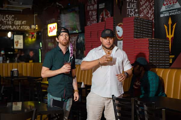 Two friends holding cocktails inside a busy pizza pub with bold wall art, booth seating, and upbeat nightlife energy.