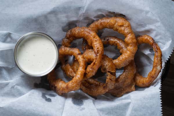 Golden-brown beer-battered onion rings served in a basket with creamy ranch dipping sauce, classic American bar appetizer on a rustic wooden table.