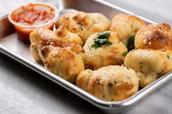 Golden-brown garlic Parmesan knots served in a metal tray with a side of marinara dipping sauce – soft, oven-baked Italian bread bites perfect as a pizzeria appetizer.
