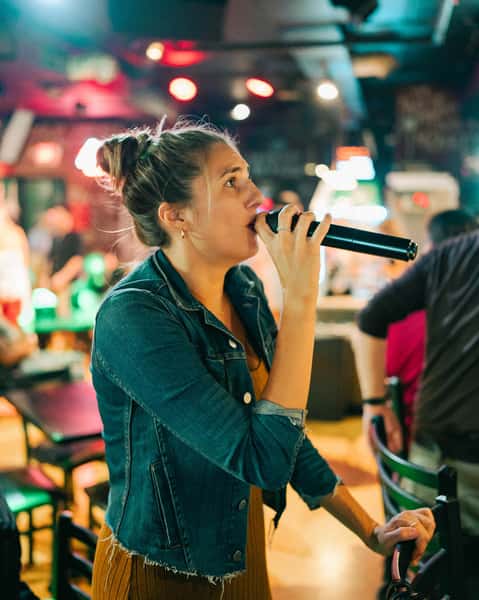Woman singing karaoke into a microphone inside a vibrant pizza and sports bar with colorful lights and energetic nightlife atmosphere.