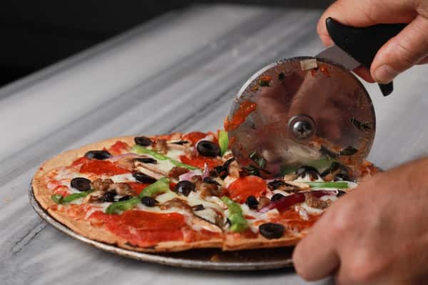 Thin-crust veggie pizza topped with olives, mushrooms, green peppers, and pepperoni being sliced with a pizza cutter in a pizzeria kitchen.