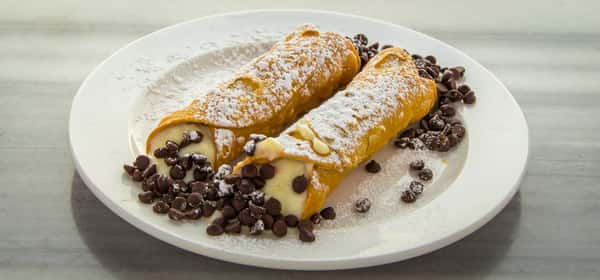 Two classic Italian cannoli filled with sweet ricotta cream, dusted with powdered sugar, and topped with chocolate chips at a pizza and dessert spot.