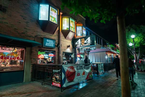 Nighttime exterior of a bustling pizza pub with neon signs, patio seating, and illuminated storefront in a downtown entertainment district.