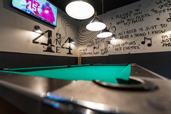 Modern sports bar interior featuring a green felt pool table, pendant lighting, wall mural art, and mounted TV creating a lively entertainment atmosphere.