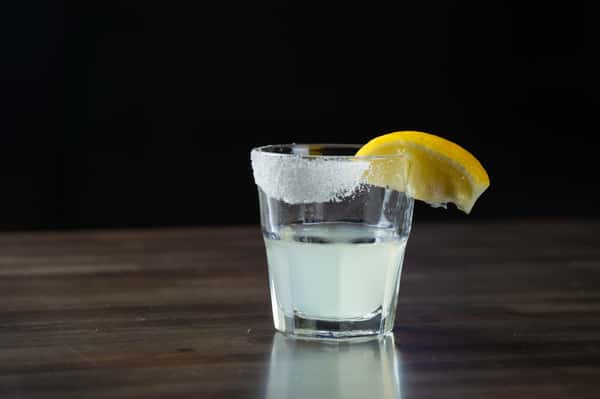 Chilled lemon drop shot with a sugar-rimmed glass and fresh lemon wedge garnish, served on a dark wooden bar top – refreshing citrus cocktail perfect for a lively bar or upscale restaurant lounge.