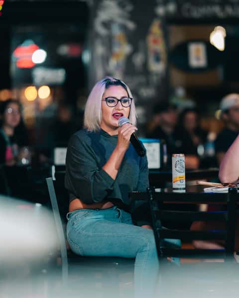 Woman singing karaoke at a table inside a vibrant neighborhood bar and grill, with craft beer can nearby and blurred crowd in the background.
