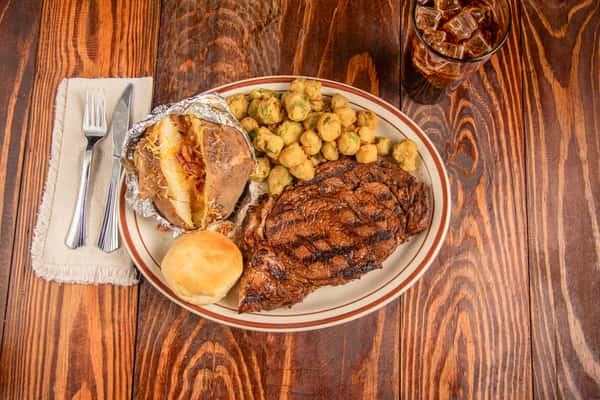 Top view of grilled ribeye steak with baked potato, roll, and seasoned sides at Blackwell Revival in Atkins, AR