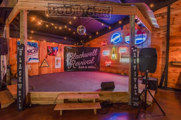 Live music stage with rustic wood backdrop and neon signs at Blackwell Revival in Atkins, AR, showcasing a lively Cajun-inspired atmosphere
