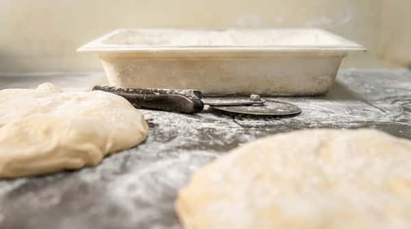 Two mounds of dough on a flour-dusted surface with a pizza cutter and a flour-covered container.