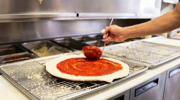Hand spreading red tomato sauce on raw pizza dough with a ladle on a metal grate in a kitchen.