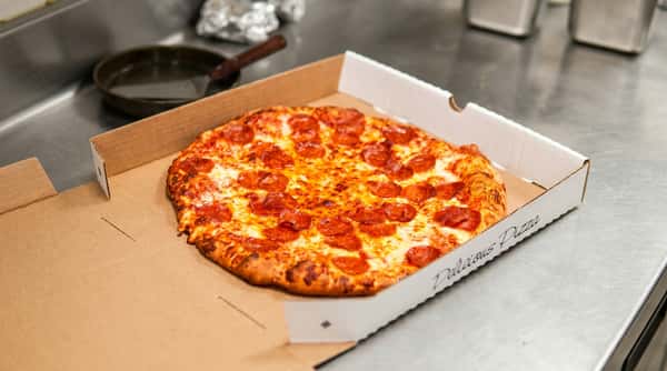Pepperoni pizza in an open cardboard box on a stainless steel counter, with "Delicious Pizza" visible on the box.