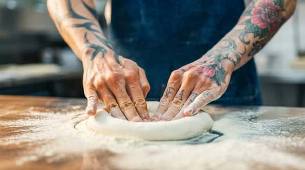 Tattooed hands covered in flour pressing down on a round of dough on a floured surface.