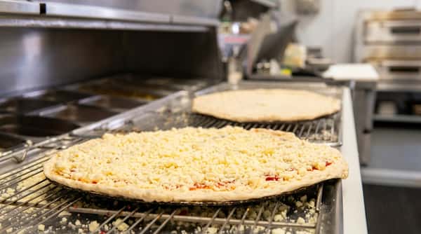 Uncooked pizza with shredded white cheese on a metal conveyor belt; hands prepare another pizza in the background.