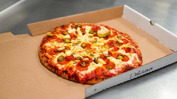 Pepperoni pizza with pineapple and jalapeños in an open cardboard box on a metal surface.