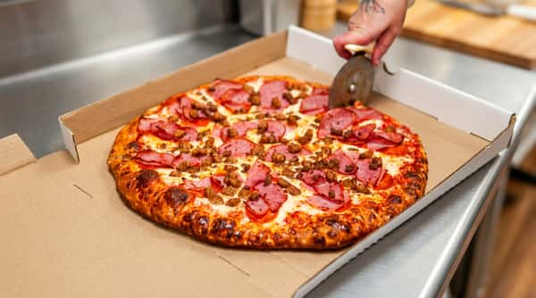 Pizza in a box being sliced, topped with pepperoni, ham, and crumbled sausage.