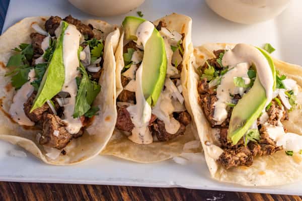 Three grilled steak tacos on flour tortillas, topped with avocado slices, onions, cilantro, and creamy chipotle sauce.