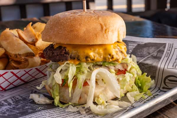 Juicy cheeseburger with shredded lettuce and tomato on a toasted bun, served with seasoned potato wedges in a red checkered tray.