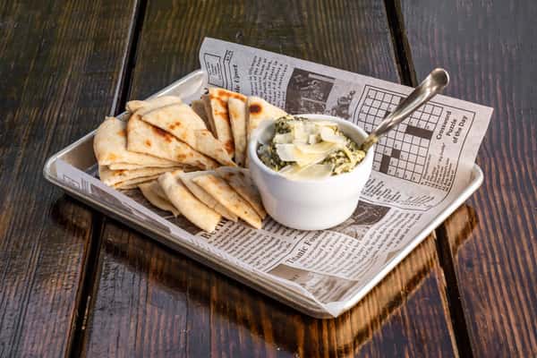 Spinach artichoke dip topped with shaved Parmesan, served with warm pita wedges on a newspaper-lined tray.