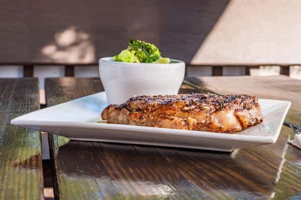 Grilled peppered steak served on a square white plate with a side of steamed broccoli and squash in a small white bowl.