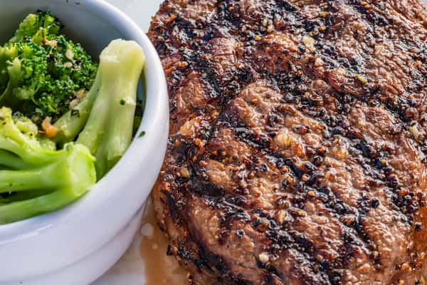 Close-up of a juicy grilled steak with cracked pepper seasoning, served alongside steamed broccoli in a white ceramic bowl.