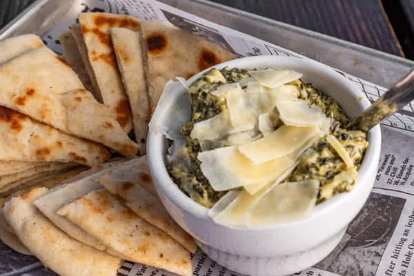 Creamy spinach artichoke dip topped with shaved Parmesan, served with warm grilled pita wedges on a metal tray.