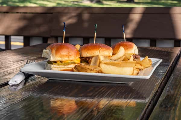 Trio of cheeseburger sliders with pickles and a side of thick-cut potato wedges served on a white plate at an outdoor table.