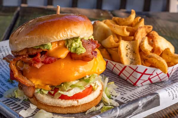 Cheeseburger stacked with bacon, cheddar, guacamole, lettuce, and tomato, served with seasoned curly fries on a metal tray.