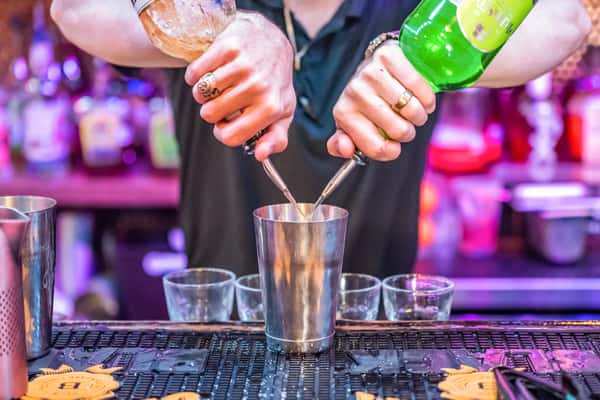 Bartender pouring two different liquors into a cocktail shaker at a vibrant, neon-lit bar counter.