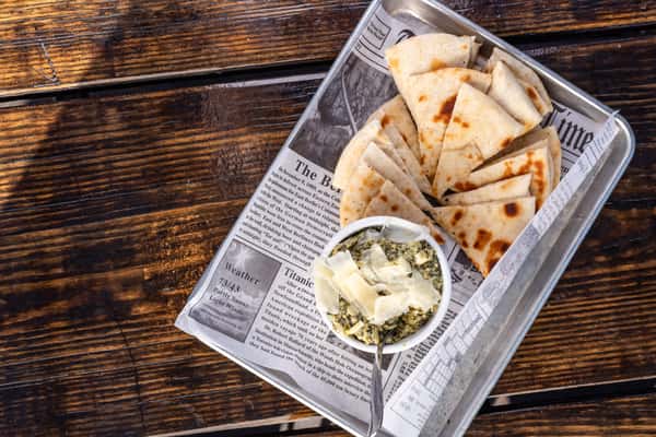 Warm grilled pita wedges served with creamy spinach artichoke dip topped with shaved Parmesan on a newspaper-lined metal tray.