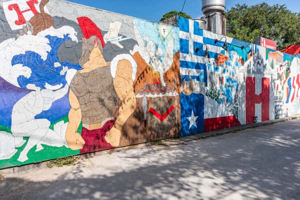 This colorful mural blends Greek mythology and culture with Texas pride. It features a Spartan warrior, Greek architecture, doves, waves, and symbols from both Greece and Texas—including the Greek flag and the Texas Lone Star—alongside the "H" logo for Humphrey's, tying the artwork to the restaurant it decorates.