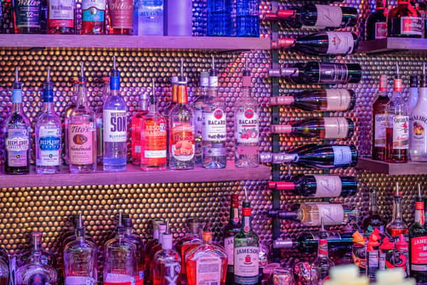 Colorful bar display with shelves stocked with vodka, rum, whiskey, liqueurs, and red wine bottles under moody purple lighting.