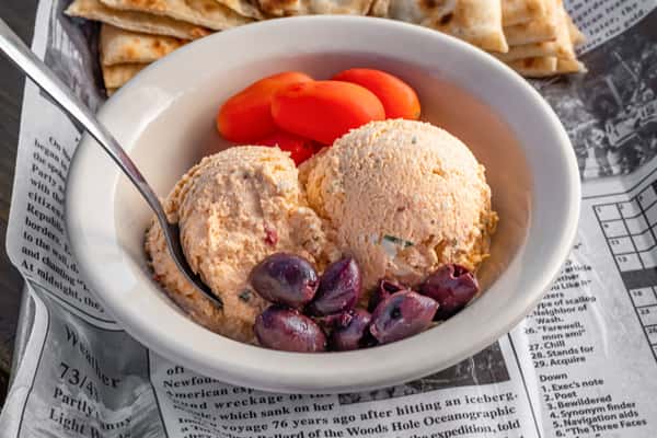 Creamy whipped feta dip with kalamata olives and grape tomatoes, served with warm pita bread on a newspaper-lined tray.