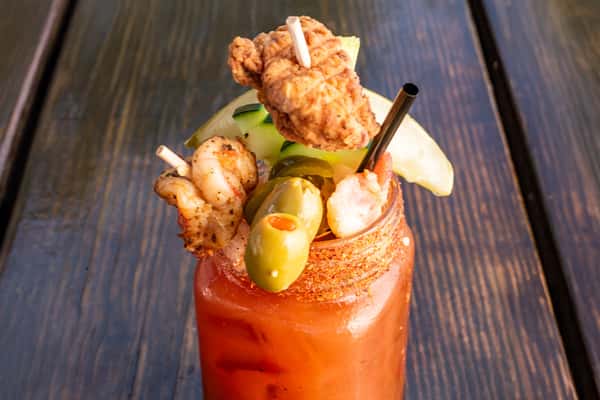 Spicy Bloody Mary in a mason jar rimmed with seasoning, garnished with fried chicken, shrimp, olives, cucumber, and lime.