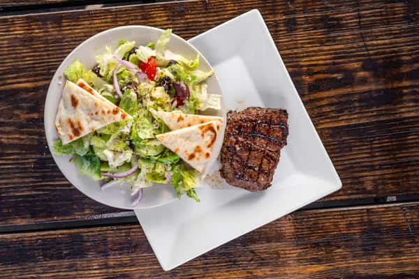 Grilled steak served with a fresh Greek salad topped with kalamata olives, red onions, and pita wedges on a white plate.