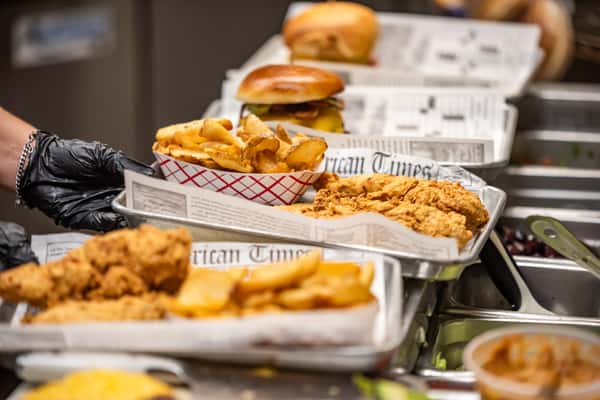 Assorted diner-style meals with fried chicken tenders, seasoned fries, and cheeseburgers served on metal trays lined with faux newspaper.
