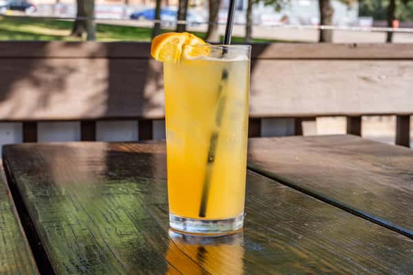 Refreshing orange cocktail served over ice in a tall glass, garnished with an orange wedge and a black straw on an outdoor table.