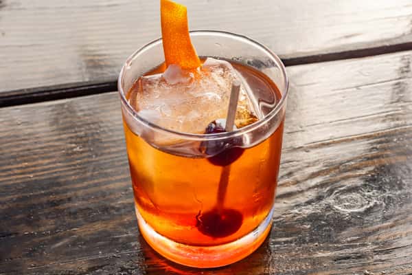 Classic Old Fashioned cocktail with a large ice cube, garnished with an orange peel and cocktail cherry in a short glass.