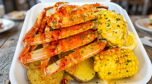 Boiled crab legs with seasoning, corn on the cob, and potatoes in a white take-out container.