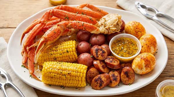 A take-out container filled with snow crab legs, corn on the cob, sliced sausage, shrimp, and a small sauce cup.