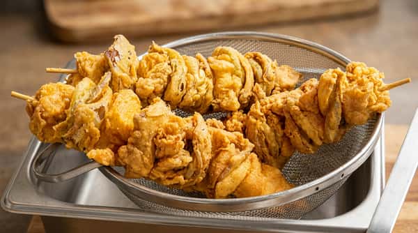 Skewered fried mushrooms or vegetables, battered and golden brown, resting in a metal strainer.