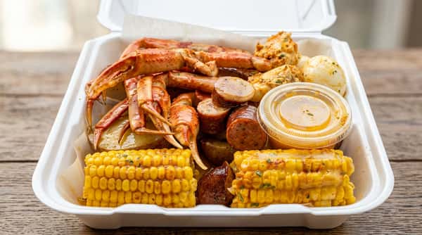Seafood boil with crab legs, sausage, corn, potatoes, and a side of butter sauce in a take-out container.