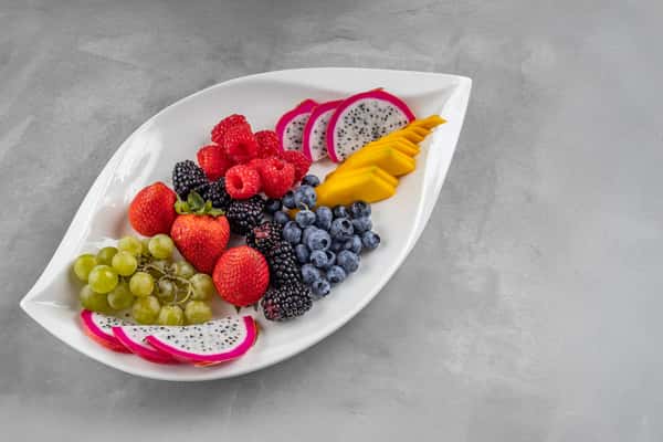 Fresh tropical fruit platter arranged on a white leaf-shaped dish, showcasing sliced dragon fruit, ripe mango, strawberries, blueberries, raspberries, blackberries, green grapes, and red grapes for a vibrant and refreshing brunch option.